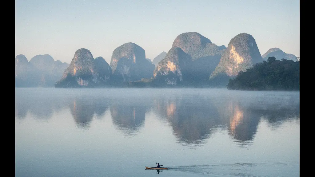 Vue panoramique du lac au lever du jour avec une brume légère, eau calme et montagnes karstiques, un petit kayak isolé au loin.